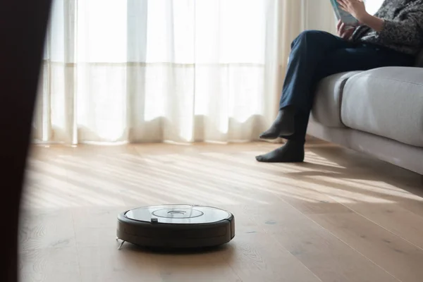 Robot elektrikli süpürge oturma odasını ahşap zeminle temizlerken bir kişi kanepede oturuyor.