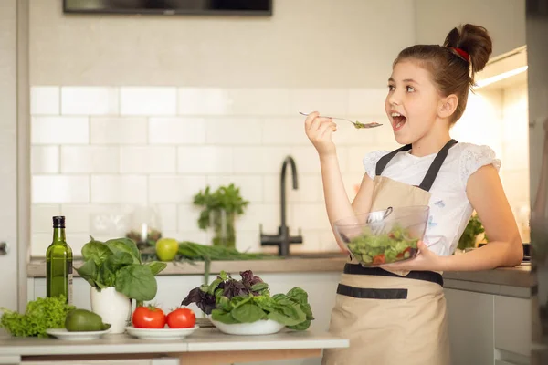 Önlüklü genç bir kız ev mutfağında tek başına salata hazırladı. Elinde bir kase pişmiş salata tutuyor ve tadına bakıyor..