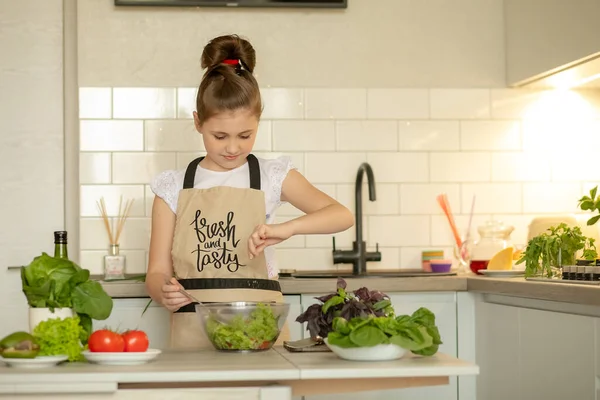 Bir kız ev mutfağında yeşil sebze salatası pişiriyor. Annemin yardımcısı pişmiş salatayı tuzladı. Kız güzel bir önlük giyiyor. Sağlıklı yaşam tarzı kavramı.
