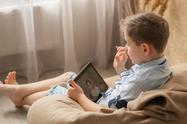 Oğlan kız arkadaşıyla internetten iletişim kuruyor. Tabletin ekranındaki kız gülümsüyor ve bir arkadaşının hikayesini dikkatle dinliyor. Online. Çocuklar evde karantinaya alındı. Bilgisayar İletişimi