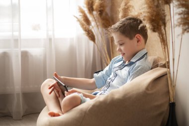 Adam internetten iletişim kuruyor. Karantina gerçeklikleri. Bilgisayar aracılığıyla iletişim. Çocuk elinde bir tablet tutuyor ve ekranla iletişim kuruyor. Çocuk rahat bir sandalyede oturur ve arkadaşlarıyla bir konferans düzenler..