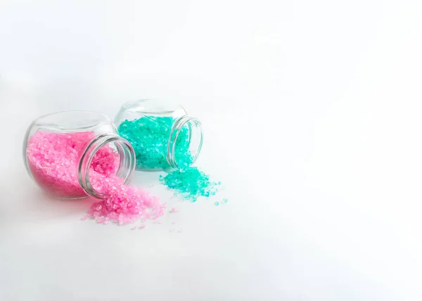 Çerçevenin sol tarafında turkuaz ve açık pembe banyo tuzu bulunan kavanozlar var. Yatay fotoğraf. Beyaz arka planda nesneler. Bankalar yan yatar. Banyo tuzu arka plana döküldü..