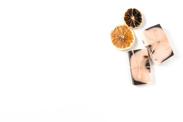 Düz kompozisyon, iki el yapımı kahverengi pembe sabun, turuncu renkli ve kuru portakal dilimli ahşap desenli beyaz arka planda. Çerçevenin sağ tarafında bulunan nesneler