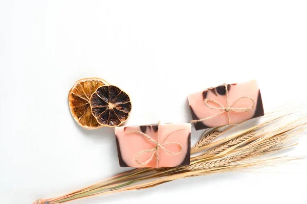 Düz kompozisyon, iki el yapımı kahverengi pembe pembe sabun, turuncu renkli, kuru portakal dilimleri ve beyaz arka planda tahta desenli bir buğday. Bulunan nesneler