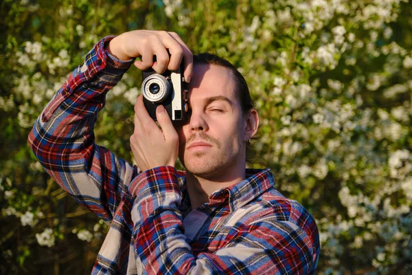 Hipster in flannel shirt is photographing flowering trees with his film soviet camera in May.