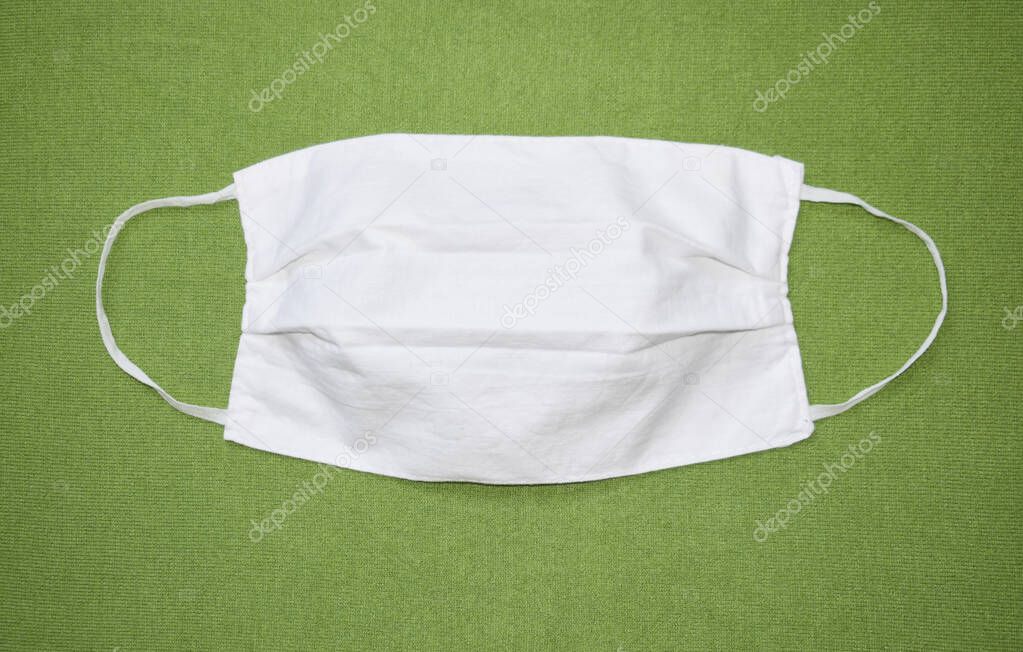 White protective face mask made of fabric on a green background. Mask with elastic bands on the sides. View from above.