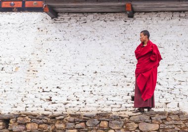 Thimphu / Bhutan - 28 Şubat 2016: Himalaya dağlarındaki manastırda beyaza boyanmış taş duvarın arkasındaki geleneksel kırmızı elbiseli genç Budist rahip..