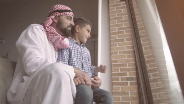 Ευτυχισμένος πατέρας και γιος στέκονται κοντά στο παράθυρο ενώ ο πατέρας δείχνει μακριά και χαμογελώντας. Αραβική έννοια της οικογένειας. — Αρχείο Βίντεο