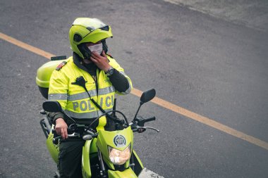 Motosikletli Kolombiyalı polis memuru sokakta maske takıyor. Yolun kenarında durmuş, yeşil güvenlik yansıtıcı üniforma ve miğfer giyiyor. Bogota, Kolombiya. 