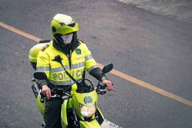 Motosikletli Kolombiyalı polis memuru sokakta maske takıyor. Yolun kenarında durmuş, yeşil güvenlik yansıtıcı üniforma ve miğfer giyiyor. Bogota, Kolombiya. 