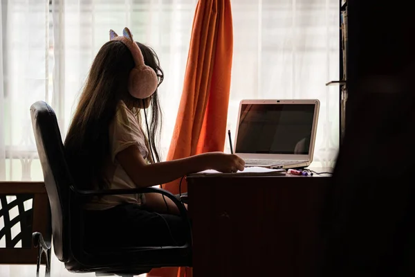 Teen girl doing a homework with computer, notebook pencils and pens at her desk; she\'s at home, indoors. She\'s wearing headphones with cat\'s ears and is beside the window. Bogota, Colombia.