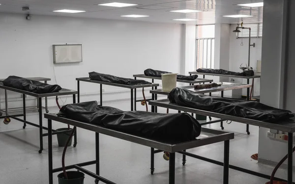 Covered human corpses on tables in a morgue / mortuary waiting for identification, autopsy, burial or cremation. Taken in Armenia, Colombia. The bodies are in a big, lonely room; they are covered with black blankets; might be the victims of pandemic.