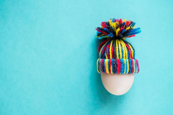 Easter card - white egg in knitted wool colored hat on blue background. Copy space for text — Stock Photo, Image