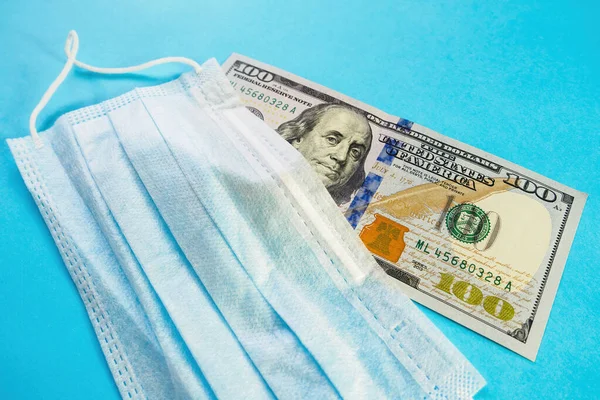 Yüz maskesi mavi arka planda 100 dolarlık banknotun üzerinde yatıyor. Tıbbi maske fiyat artışı konsepti, koronavirüse karşı koruma, grip, alerji. Boşluğu kopyala