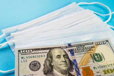 100 dolarlık banknot mavi arka plandaki tıbbi maskelerin üzerinde yatıyor. Bütçe açığı, spekülasyon ve yüz maskeleri satılmış. Seçici odak