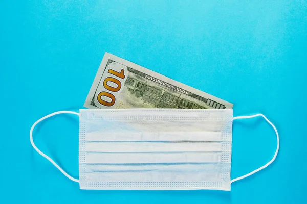 Medical mask lie on banknote of 100 dollars on blue background. Concept of deficit, speculation and sold out of face masks. Top view, copy space