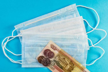 Üç tıbbi maske ve Rus parası, mavi arka planda 100 ruble ve bozuk para banknotu. Yüz maskesinin büyüme fiyatı kavramı. Üst görünüm