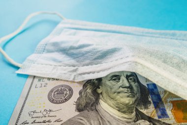 Tıbbi maske ve 100 dolarlık banknot mavi arka planda. Bütçe açığı, spekülasyon ve yüz maskeleri satılmış. Seçici odak, yakın plan