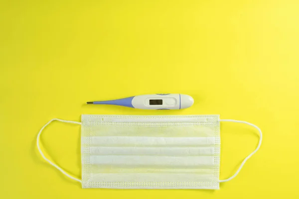 Face mask and electronic thermometer on yellow background. Top view, flat lay, copy space