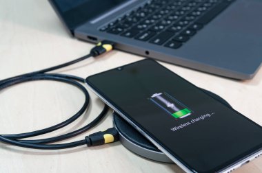 Modern akıllı telefon, kablosuz şarj cihazından şarj oluyor. Laptopun yanında, ahşap bir masanın üzerinde. Modern teknoloji konsepti. Seçici odak