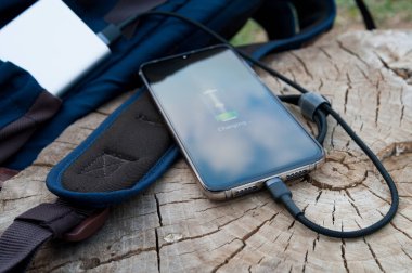 Modern akıllı telefonlar elektrik bankasından şarj oluyor. Sırt çantasının yanında tahta bir kütük var. Modern teknoloji konsepti. Seçici odak