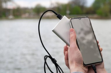 Adam nehir kıyısındaki elektrik bankasından şarj olurken beyaz ekranlı akıllı telefon kullanıyor. Modern teknoloji konsepti. Seçici odak.