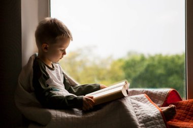 Pencerenin pervazında oturmuş kitap okuyan sevimli küçük çocuk..