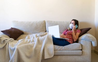 Kanepede oturan bir kadın dizüstü bilgisayar izliyor ve telefonda maskeyle konuşuyor. Karantina hayat tarzı..