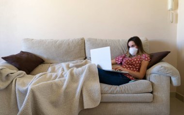 Kanepede oturan bir kadın dizüstü bilgisayar seyrediyor, internetten öğreniyor ve maskeyle evden telgraf çekiyor. Hayat tarzını karantinaya alıyor..