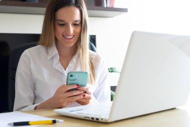 Evde dizüstü bilgisayarla çalışırken cep telefonundaki e-postaları okuyan gülümseyen genç bir girişimci. Evden taşınma yaşam tarzı.