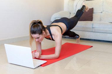 Çekici genç esmer evde egzersiz yapıyor ve spor paspasının üzerinde dikilirken dizüstü bilgisayarda video izliyor. Bacak egzersizi yapıyorum..