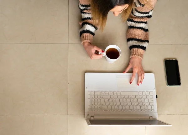 Top view of a woman working from home on the floor, typing at her laptop while holding a cup of tea. Telecommuting lifestyle.