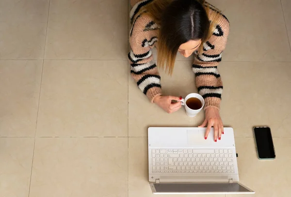 Top view of a woman working from home on the floor, typing at her laptop while holding a cup of tea.