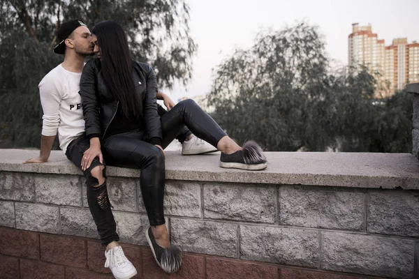couple in love: a beautiful young girl in black clothes smiling hugging a stylish young man sitting on a concrete parapet in a park on a background of tall buildings in the distance