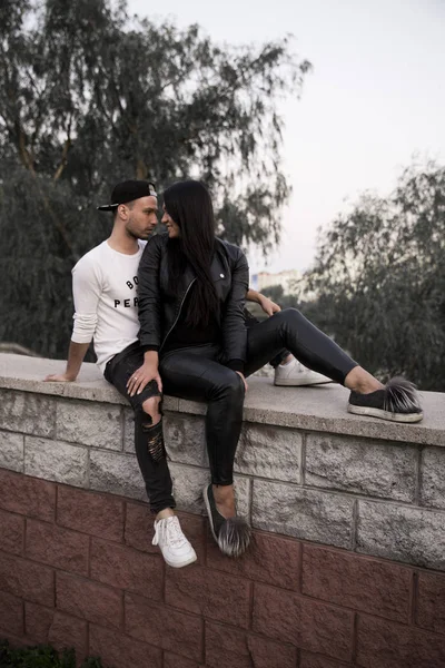 couple in love: a beautiful young girl in black clothes smiling hugging a stylish young man sitting on a concrete parapet in a park on a background of tall buildings in the distance