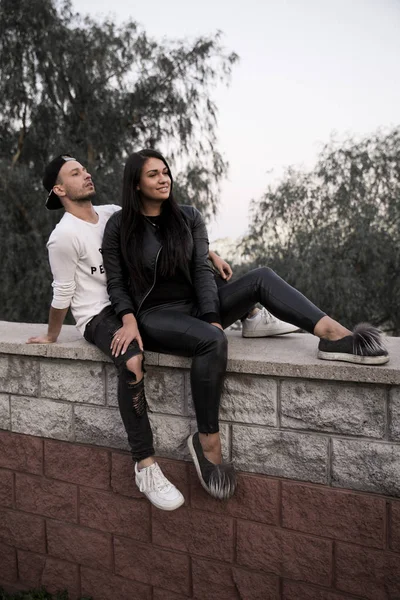 couple in love: a beautiful young girl in black clothes smiling hugging a stylish young man sitting on a concrete parapet in a park on a background of tall buildings in the distance