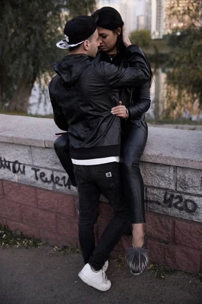 Couple in love: a beautiful young girl in a black leather jacket and pants gently hugs a stylish young man sitting on a concrete parapet in a park against a backdrop of tall buildings in the distance