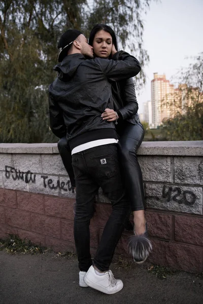 Couple in love: a beautiful young girl in a black leather jacket and pants gently hugs a stylish young man sitting on a concrete parapet in a park against a backdrop of tall buildings in the distance