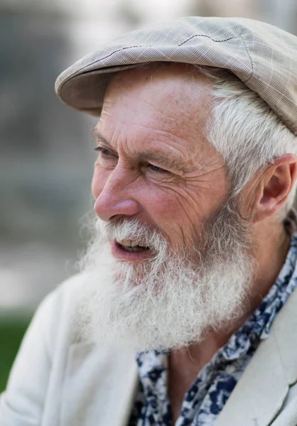 in the profile of the emotion of an expressive gray-haired old man with a beautiful strong-willed tanned face and a white beard in a light cap, blue shirt and jacket on a blurred street background