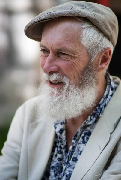 in the profile of the emotion of an expressive gray-haired old man with a beautiful strong-willed tanned face and a white beard in a light cap, blue shirt and jacket on a blurred street background