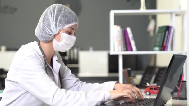 Жінка-лікар з маскою працює над чимось з комп'ютером і звертає увагу в офісі або лікарні . — стокове відео