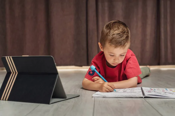 Un niño pequeño escribe en un cuaderno usando una tableta — Foto de Stock