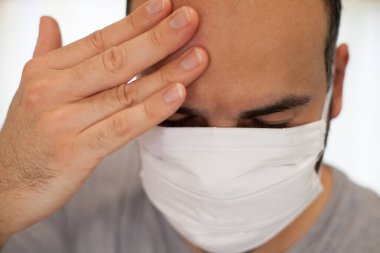 Coronavirus hastası bir adam maske takar ve ateş, yorgunluk ve baş ağrısı gibi semptomlardan dolayı alnına dokunur..