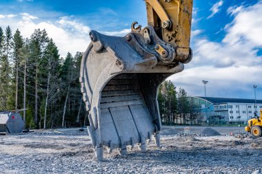 Henüz hazır olmayan yeni bir yolda büyük bir kazı kazıcının kepçesinin yan görüntüsünü kapatın. Koyu mavi bulutlarla güneşli bir akşam, bahar zamanı, karışık orman ve arka planda eski kapalı hokey stadyumu..