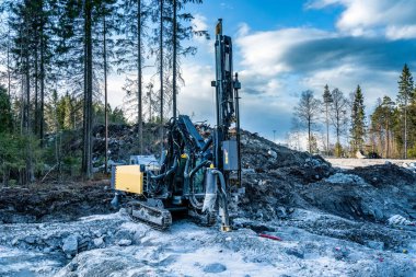 Granit kayaların üzerinde duran sarı sondaj makinesinin ön görüntüsü ve kaya patlatma alanındaki dinamit için sıkıcı delikler - daha fazla kaya patlaması, yeni yol inşaatı. Akşam gölgeleri, yeşil orman