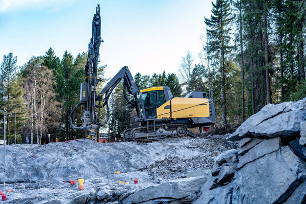 Side view at yellow drilling machine which stands on granite rock and boring holes for dynamite at rock blasting area - further rock exploding, new road construction. Evening shadows, green forest