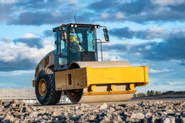 Eye catching yellow road roller with enclosed climate controlled cabin stands on not ready new road, stones, blue sky, clouds, front left side view. Clean shiny old heavy tractor clipart