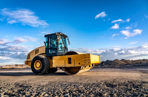 Eye catching yellow road roller with enclosed climate controlled cabin stands on not ready new road, stones, blue sky, clouds, left side view. Clean shiny old heavy tractor