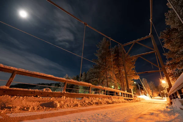 Night view at the Moon above the metal cable-stayed bridge in Swedish countryside, it illuminated by lights, cold winter blue skies with stars and thin clouds. Lappland, Northern Sweden