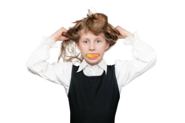 Liseli kız, ağzında bir elmayla yüzünü buruşturuyor ve beyaz arka planda saçlarını kafasında parçalıyor.